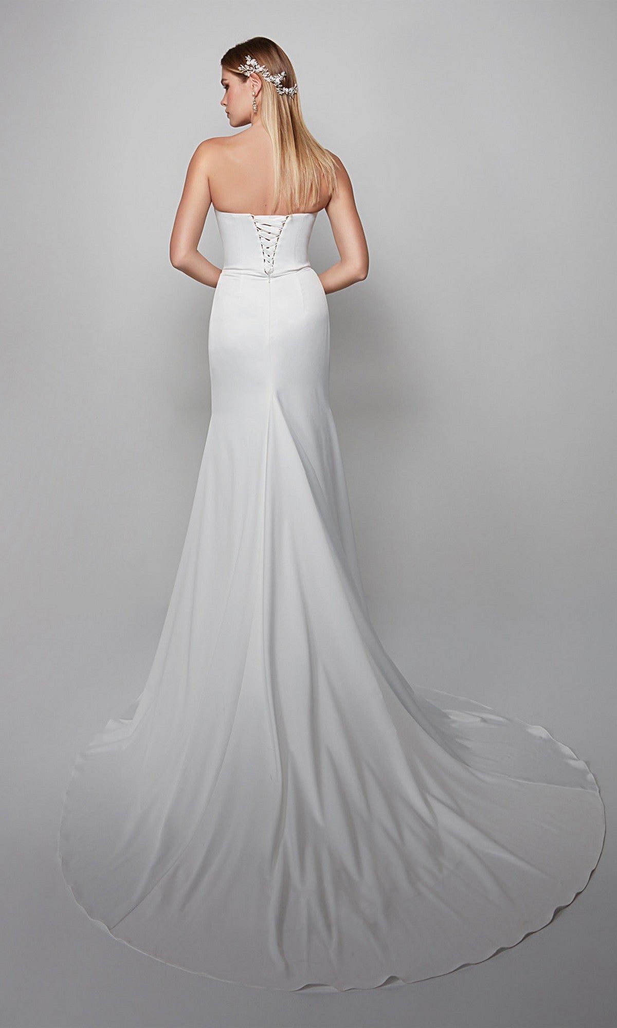 A woman faces away, wearing the Alyce 7056 long formal dress—strapless, white with a sweetheart neckline, lace-up back, and a sweeping train spread on the floor.